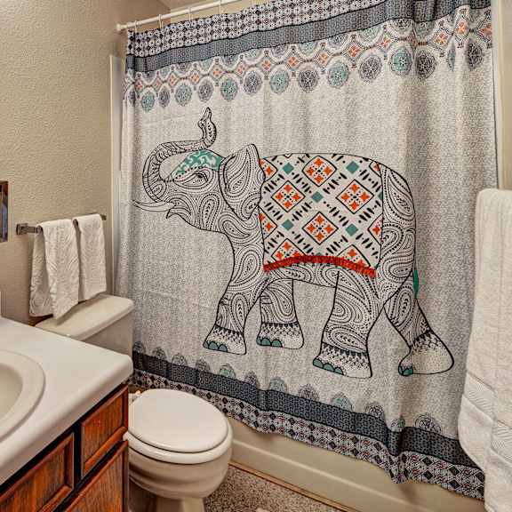 a bathroom here at bay arms apartments with a patterned elephant shower curtain. features a white sink, wooden cabinets, a mirror, and neatly hung white towels, conveying a cozy atmosphere.