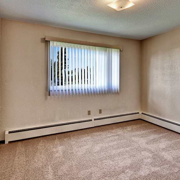 empty beige bedroom here at bay arms apartments with carpeted floor, a bright window with vertical blinds, and baseboard heating. soft overhead light creates a calm atmosphere.