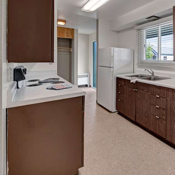 a modern kitchen here at bay arms apartments with brown wooden cabinets, white countertops, and a white refrigerator. the room is well-lit with a window above the sink.