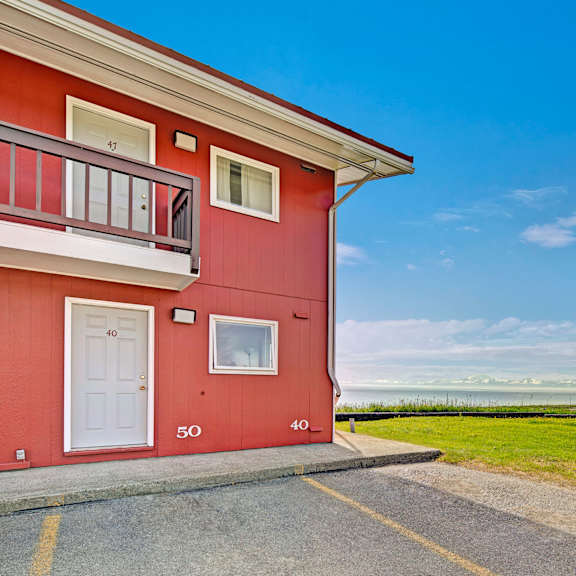 our red building at bay arms apartments with white doors, numbers 40 and 50 visible. expansive view of grassy landscape, ocean, distant snow-capped mountains, under a clear blue sky.