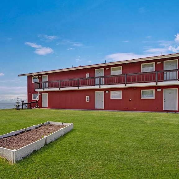 our red barn-style building here at bay arms apartments with multiple doors and windows sits on a lush green lawn. a white raised garden bed lies in front. the sky is clear and blue.