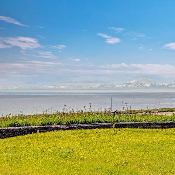 wide landscape view from bay arms apartments of a grassy field leading to a sandy shore, with a calm ocean and distant snow-capped mountains under a clear blue sky.