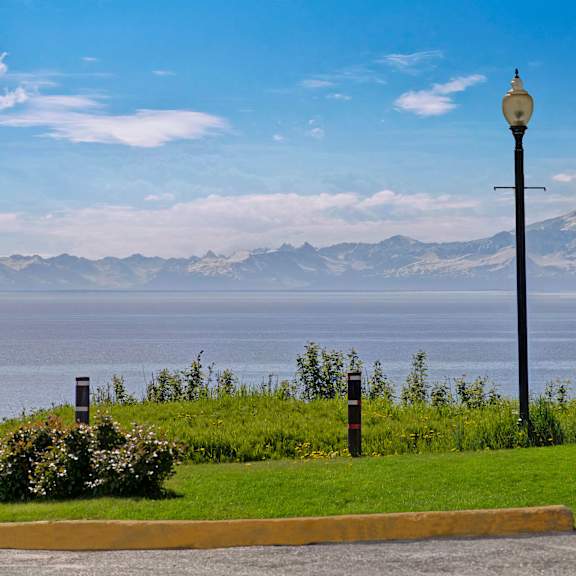 a serene coastal view from bay arms apartments with a lamp post and green grass in the foreground, a calm sea, a distant ship, and snow-capped mountains under a clear blue sky.