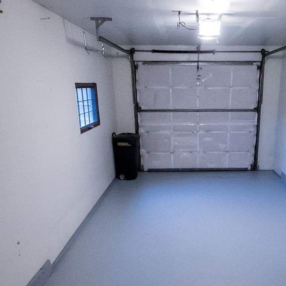 empty garage here at birchwood apartments with white walls and light gray floor, bright overhead light, closed garage door at the back, and a small window on the left. minimalistic and clean.