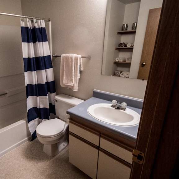 compact bathroom here at birchwood apartments with a blue and white striped shower curtain, a sink with a blue countertop, toilet, towel rack, and shelf with folded towels. clean and neat.