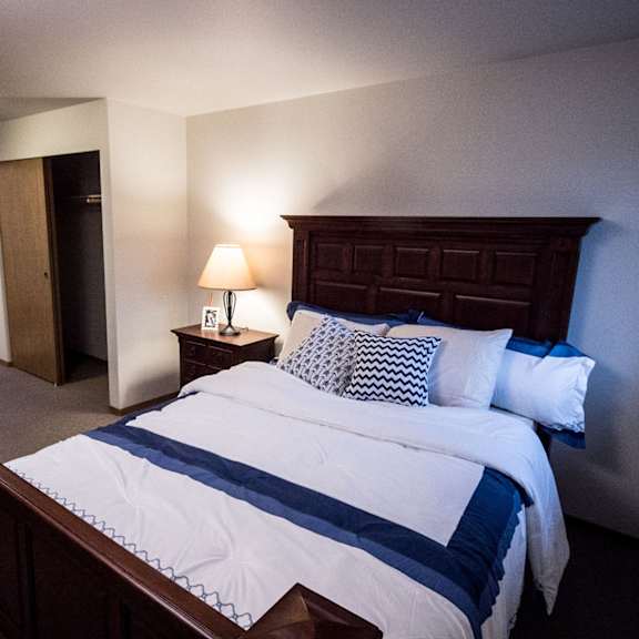 cozy bedroom here at birchwood apartments with a dark wood bed frame, white and blue bedding, and patterned pillows. a lit lamp on a bedside table adds warmth. closet door ajar.