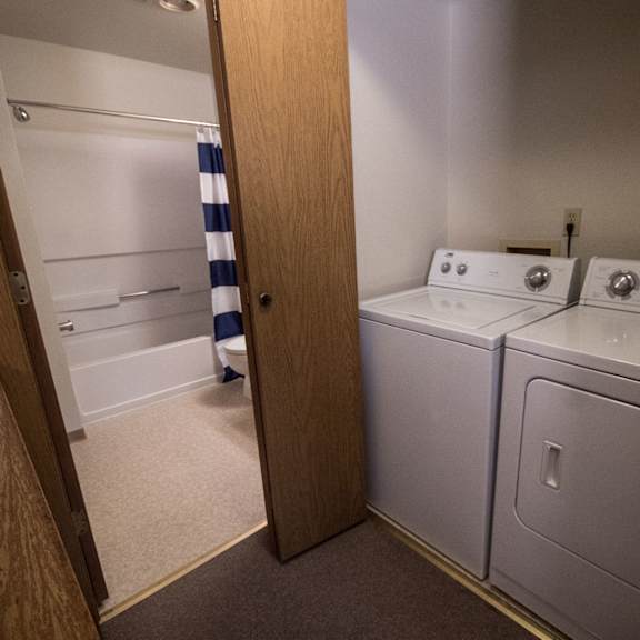 compact laundry room here at birchwood apartments with a washer and dryer next to a bathroom featuring a tub. a blue and white striped shower curtain adds a pop of color.