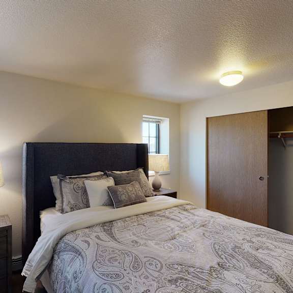 cozy bedroom here at birchwood homes apartments with a large bed, patterned bedding, and two lamps on nightstands. natural light filters through small windows, enhancing the calm ambiance.