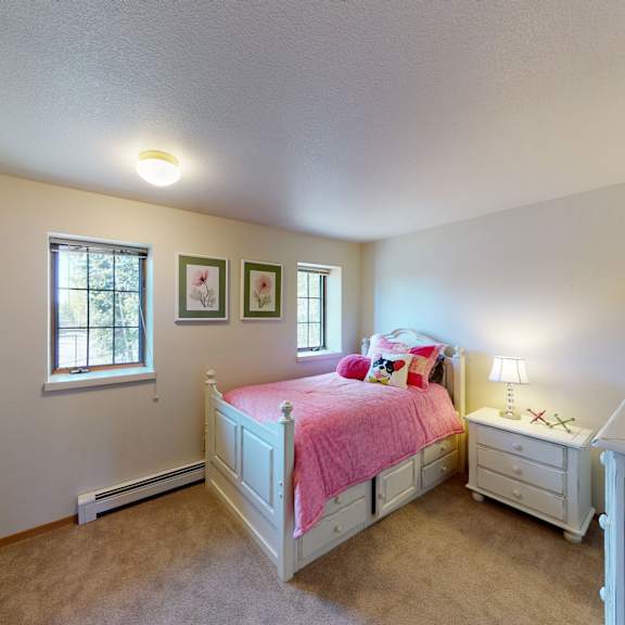cozy bedroom here at birchwood homes apartments with a white daybed covered in a pink comforter and colorful pillows. soft lighting from a lamp. two floral pictures on the wall.