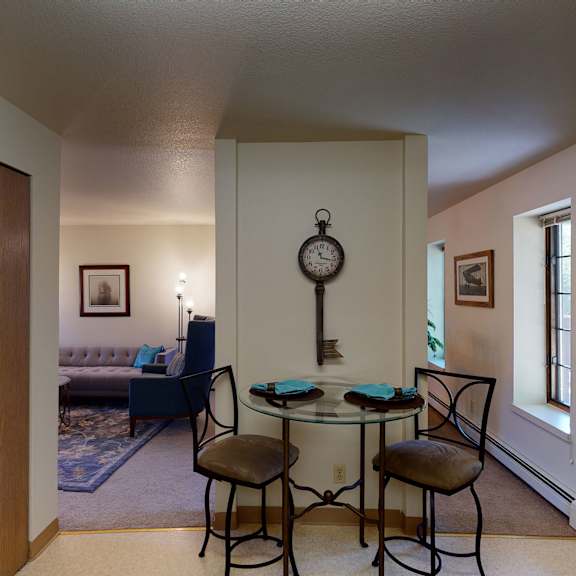 cozy room here at birchwood apartments with a small dining nook featuring a round glass table and two chairs. a large key-shaped clock hangs on the wall. a living area with a sofa and framed art is visible in the background.