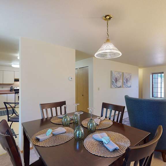 dining room view of a cozy home with a dark wood table set for four, elegant place settings, and a bright living area featuring plush blue chairs and large windows. birchwood homes apartments.