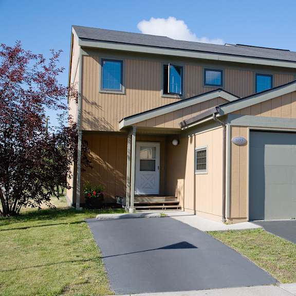 two-story beige townhouse with a one-car garage, white front door, and small porch. manicured lawn and a tree under a clear blue sky. calm suburban setting. birchwood homes.