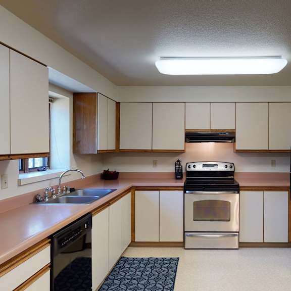 modern kitchen here at birchwood apartments with beige cabinets, stainless steel stove and refrigerator, pink countertops, and a blue patterned rug. bright overhead lighting.
