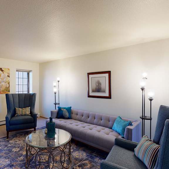 bright living room here at birchwood apartments with modern design; features a gray tufted sofa, blue armchairs, floral artwork, and floor lamps. large windows illuminate the space.