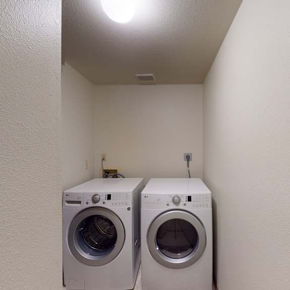 compact laundry room here at birchwood homes apartments with white walls, featuring a front-loading washer and dryer side by side under a bright ceiling light, conveying a clean, functional space.