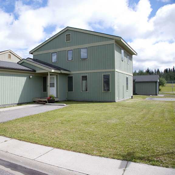 two-story green house with sloped roof, attached garage, and small porch. set in a suburban neighborhood with a grassy lawn, under a partly cloudy sky. birchwood homes.