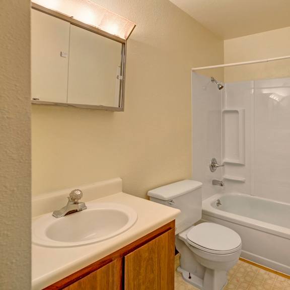compact beige bathroom here at brighton woods apartments with a white bathtub, showerhead, toilet, and sink with a mirror above. simple, clean, and functional design.