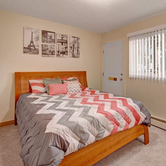 cozy bedroom here at brighton woods apartments with a wooden bed, chevron-patterned bedding in gray, white, and pink. sunlight filters through vertical blinds on a window, brightening the space. black and white art above the bed adds a modern touch.