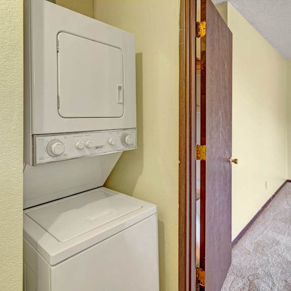 stacked washer and dryer here at brighton woods apartments in a small laundry area with beige walls. an open wooden door leads to a carpeted room, creating a cozy and tidy atmosphere.
