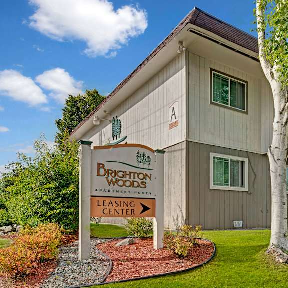 apartment complex with a beige facade and red door, surrounded by lush greenery and birch trees. a sign reads "brighton woods leasing center."
