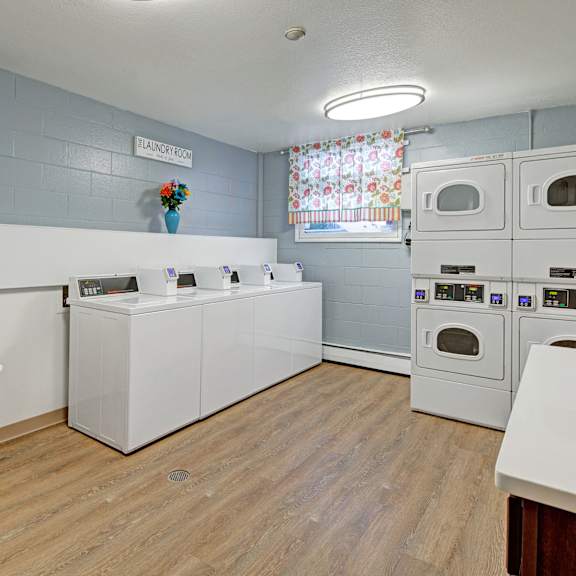 laundry room here at century plaza with a cozy feeling. features four white washers, stacked dryers, a utility sink, floral curtain, wood floor, and decorative flowers.