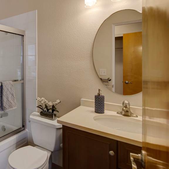 modern bathroom here at century plaza with white tiles and glass shower. a round mirror hangs above a sink with a soap dispenser. neutral tones convey a clean, calm ambiance.