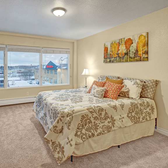 a cozy bedroom here at century plaza with beige walls and carpet, featuring a bed with patterned bedding and colorful cushions. large window shows a snowy view outside.
