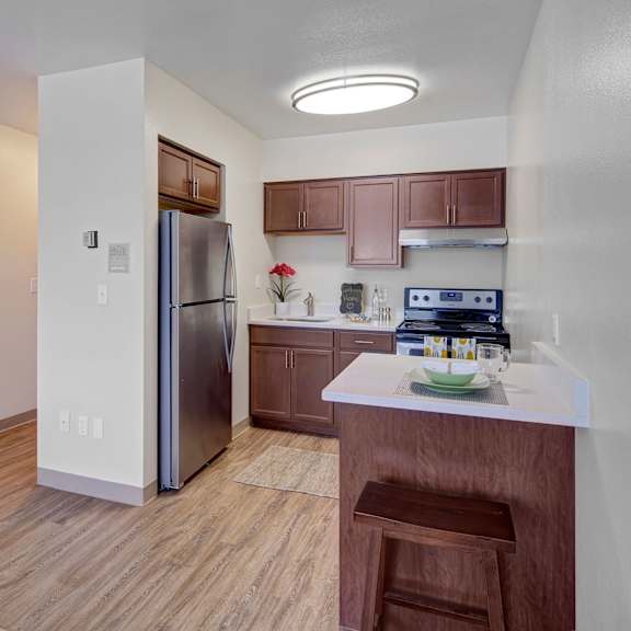 compact kitchen here at century plaza with stainless steel fridge, dark wood cabinets, and electric stove. light wood flooring and a small bar area with a stool. warm, welcoming tone.