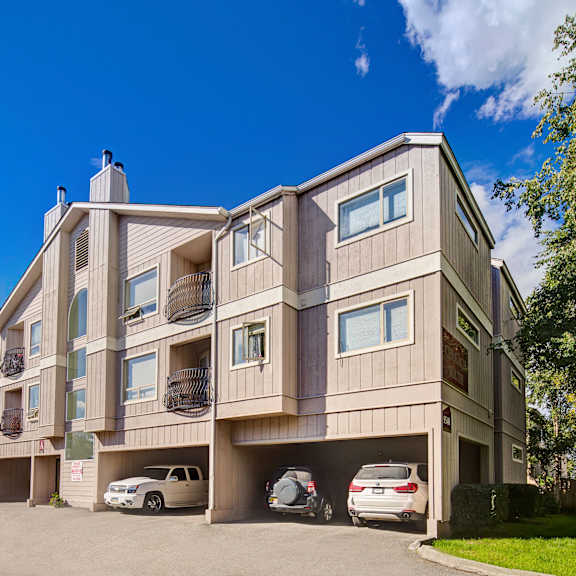 three-story beige chugach south apartment building sits under a clear blue sky, with three cars parked in open garages. trees line the right, creating a serene suburban feel.