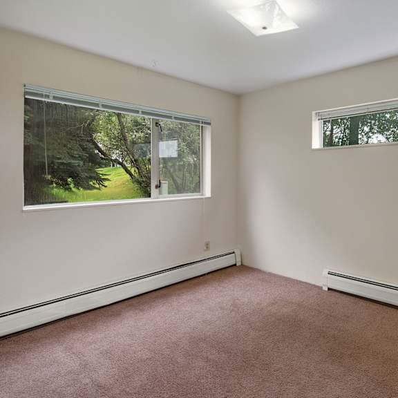 empty living room here at chugach south apartements with beige carpet and cream walls, featuring two large windows showing a green, leafy backyard. the peaceful ambiance is bright and open.