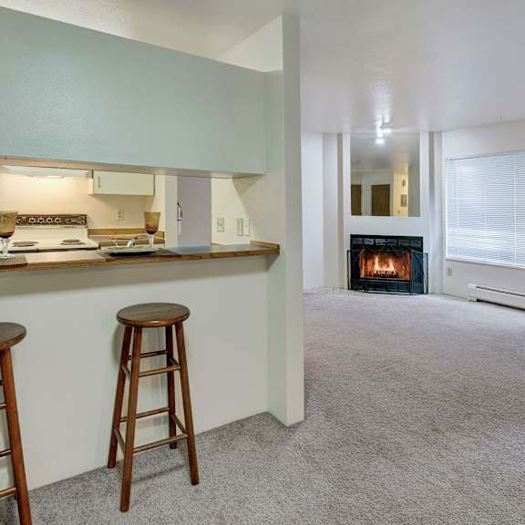 spacious, cozy living area here at chugach south apartments with a grey carpet, featuring a kitchen bar with two wooden stools on the left, and a lit fireplace on the right.