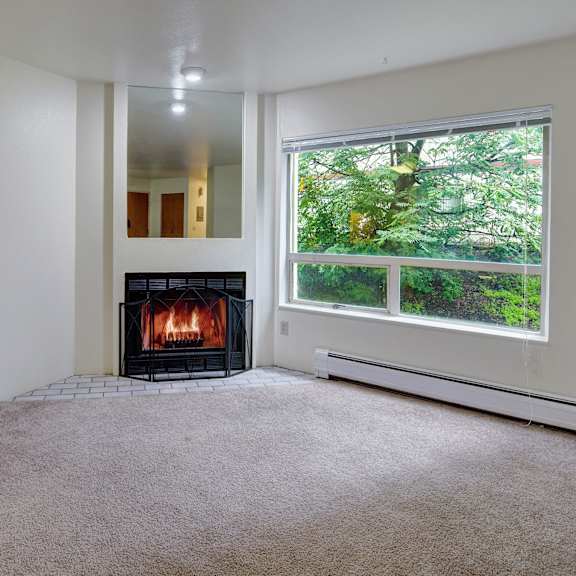 a cozy room here at chugach south apartments with a lit fireplace, beige carpet, and a large window showing green foliage. the atmosphere is warm and inviting, ideal for relaxation.