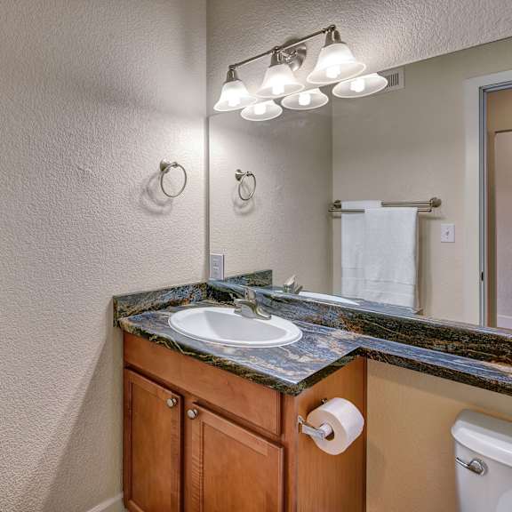 bathroom here at city view apartments with wooden vanity, marble countertop, and mirror. three-light fixture above, white towels on racks, and a roll of toilet paper. clean and modern.