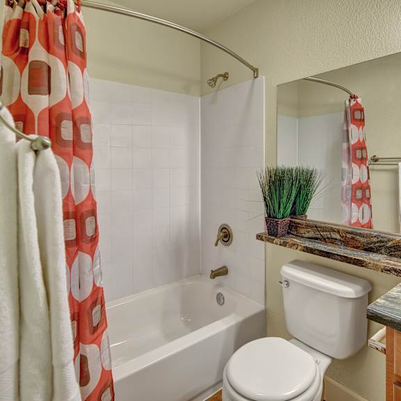 a small bathroom here at city view apartments with a white tub and colorful red and white shower curtain. a marble countertop with a sink and potted plant adds a touch of elegance.