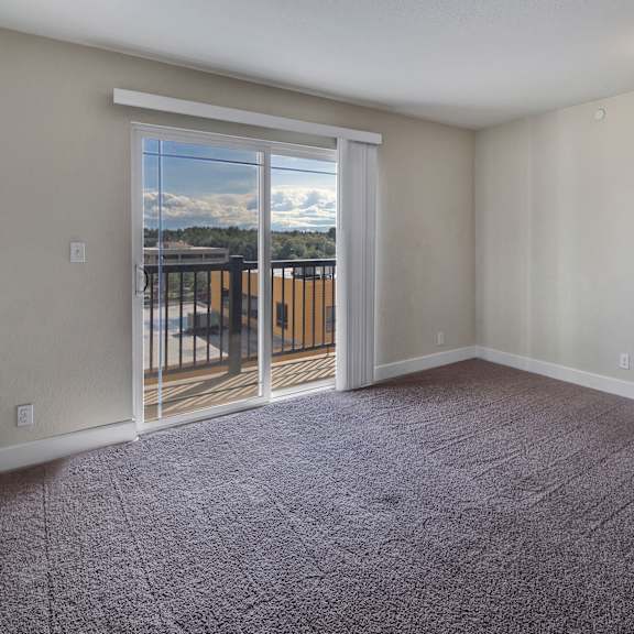 living room here at city view apartments with beige walls and gray carpet, featuring a large glass sliding door leading to a balcony. bright natural light and neutral decor.