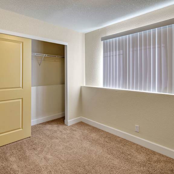 empty bedroom here at city view apartments with beige carpet, a partially open sliding yellow closet door on the left, and a bright window with vertical blinds on the right.