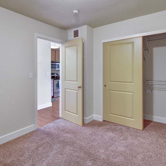 bedroom here at city view apartments with beige carpet and light walls. a partially open closet with sliding doors is on the right, and an open door leads to a kitchen.