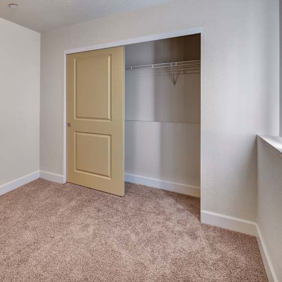 empty beige bedroom here at city view apartments with carpet, open closet with a sliding yellow door, white baseboards, and a small window on the right, evoking a clean, neutral feel.