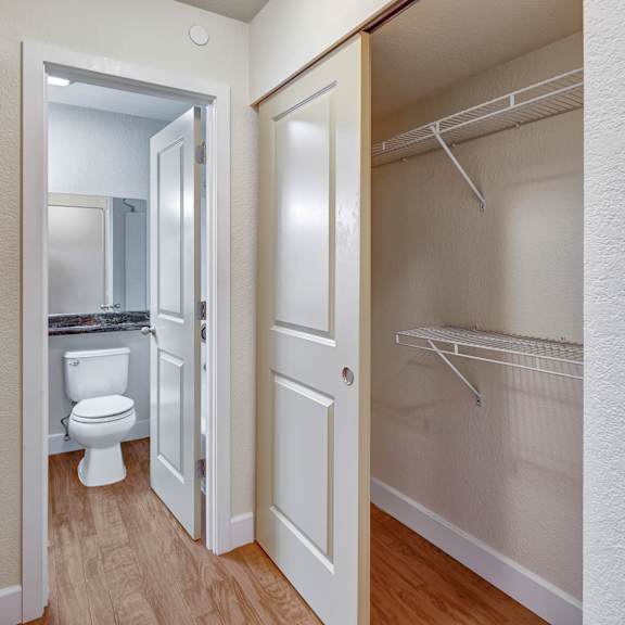 hallway here at city view apartments showing an open closet with wire shelves to the right, and a view into a bathroom with a white toilet and mirror on the left. wood flooring.