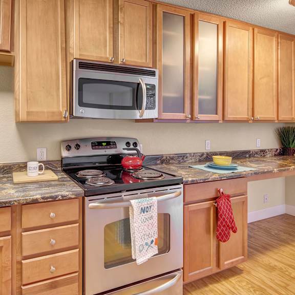 modern kitchen here at city view apartments with light wood cabinets, granite countertops, and stainless steel appliances. a red kettle sits on the stove, with oven mitts and towels nearby.