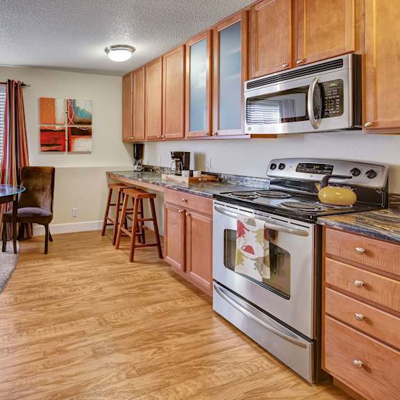 warm kitchen-dining area here at city view apartments with wooden cabinets, stainless steel appliances, round glass table, two chairs, large window, and abstract art on the wall.