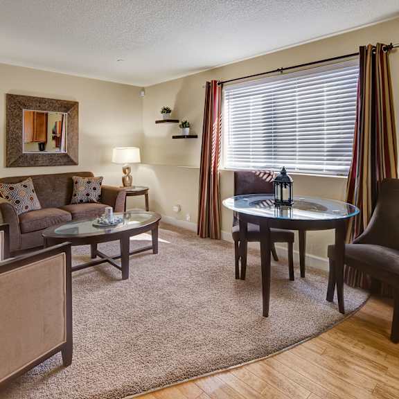 a cozy living room here at city view apartments with beige walls features a brown sofa with patterned cushions, two armchairs, and a glass coffee table. two side tables with lamps flank the sofa. a round dining table with a decorative lantern is near a window with blinds and striped curtains, creating a warm, inviting atmosphere.