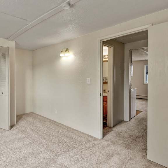empty beige carpeted bedroom here at college view apartments with an open closet on the left, showing hanging rod, and an open door leading to a hallway. dim wall light creates a cozy feel.