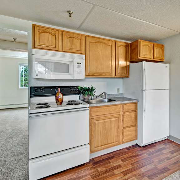 compact kitchen here at college view apartments with wooden cabinets, white appliances, and a vase on the stove. bright room with hardwood floors, adjacent to a carpeted space.