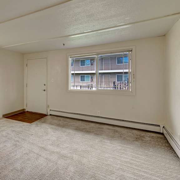 empty living room here at college view apartments with beige carpet, cream walls, and a white door. a window reveals an apartment building view. overhead light casts a soft glow, creating a neutral tone.
