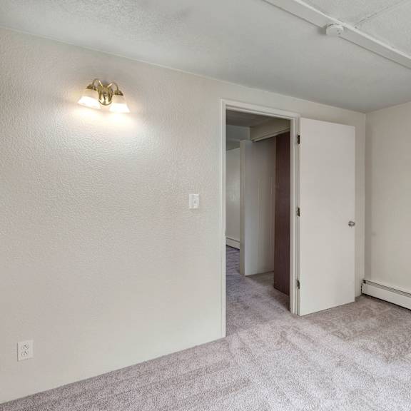empty bedroom here at college view apartments with beige walls, light gray carpet, a closed white door slightly ajar, and a wall sconce casting soft light, creating a neutral tone.