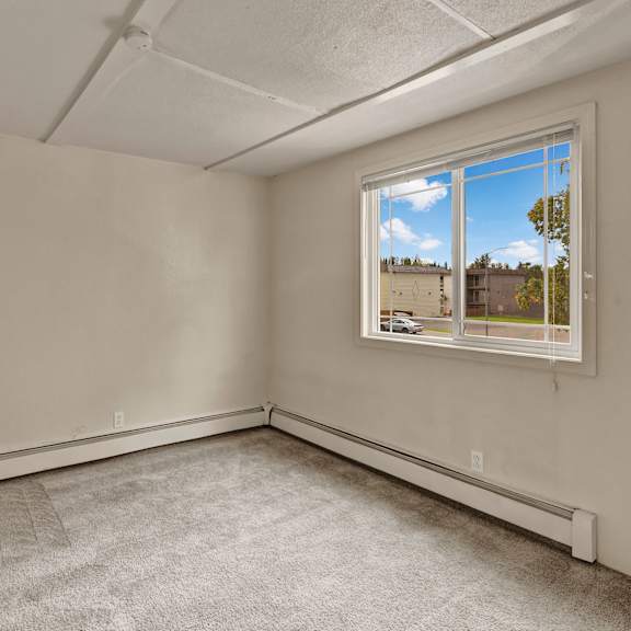 empty bedroom here at college view apartments with beige walls, light gray carpet, and a large window revealing a view of trees and buildings outside. the tone is calm and neutral.