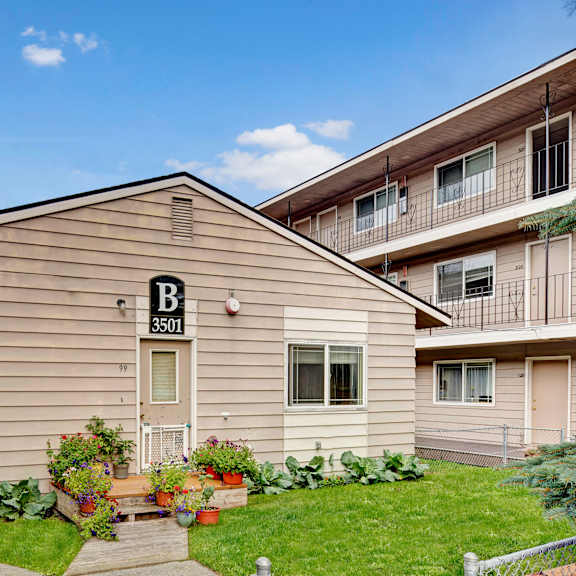 college view apartments exterior of a beige building labeled "b 3501" with potted plants and flowers in front. surrounding buildings have balconies. clear blue sky above.