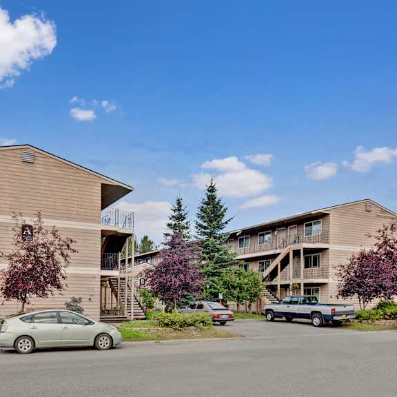 two beige apartment buildings here at college view apartments with wooden siding, surrounded by trees and parked cars under a clear blue sky. the scene conveys a peaceful suburban setting.