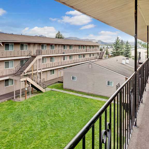 college view apartment complex with three stories, beige siding, and black railings. a green courtyard is visible below, under a partly cloudy blue sky.